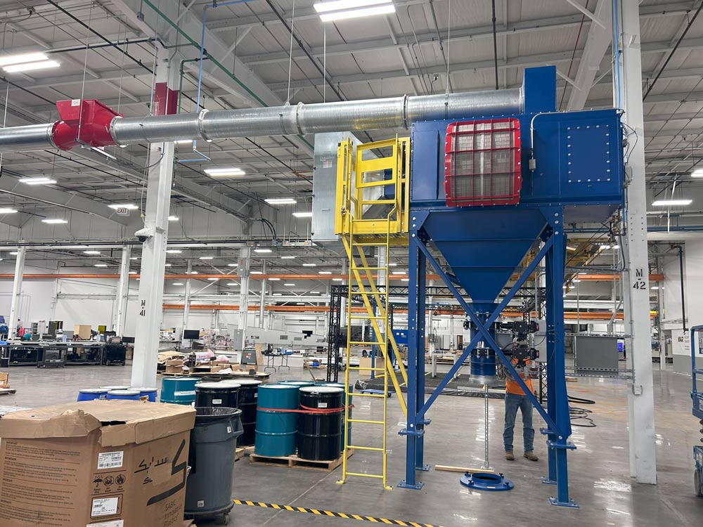 Large blue industrial dust collector with overhead ductwork installed in a manufacturing facility
