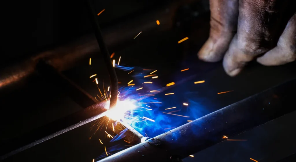 Close-up of MIG welding arc with bright blue core, orange and yellow sparks, and visible weld spatter on metal plates